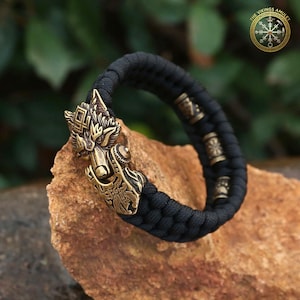 Puede incluir: Una pulsera trenzada negra con un cierre de cabeza de lobo de bronce. La pulsera tiene cuentas decorativas de bronce y est&aacute; colocada sobre una roca. El logotipo de The Vikings Amulet est&aacute; en la esquina superior derecha.