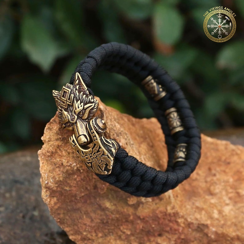 Puede incluir: Una pulsera trenzada negra con un cierre de cabeza de lobo de bronce. La pulsera tiene cuentas decorativas de bronce y est&aacute; colocada sobre una roca. El logotipo de The Vikings Amulet est&aacute; en la esquina superior derecha.