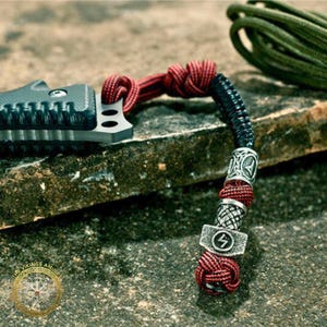 Könnte beinhalten: Ein schwarzes und silbernes Messer mit einem rot-schwarzen Schlüsselband mit dekorativen Metallperlen und einem Thorhammer-Anhänger. Das Schlüsselband ist am Griff des Messers befestigt. Im Hintergrund ist ein grünes Seil zu sehen.