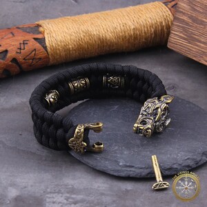 Puede incluir: Pulsera trenzada negra con detalles met&aacute;licos color bronce. La pulsera presenta un cierre de cabeza de lobo y cuentas decorativas. Tambi&eacute;n se ven un pasador de bronce y un logotipo.
