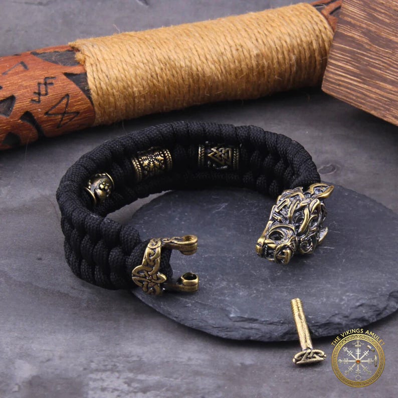 Puede incluir: Pulsera trenzada negra con detalles met&aacute;licos color bronce. La pulsera presenta un cierre de cabeza de lobo y cuentas decorativas. Tambi&eacute;n se ven un pasador de bronce y un logotipo.
