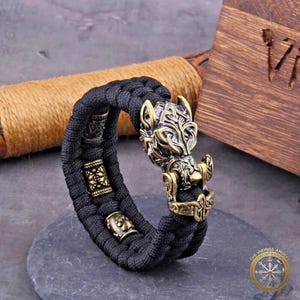 Puede incluir: Pulsera trenzada negra con cabeza de lobo de color bronce y cuentas decorativas. El cierre de la cabeza de lobo presenta detalles intrincados. La pulsera se exhibe sobre una superficie de piedra gris oscuro.