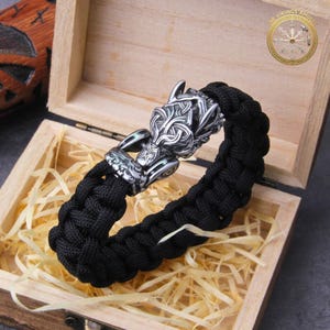 May include: Black braided bracelet with a silver-colored wolf head clasp. The bracelet is displayed in an open wooden box with straw-like filler. The wolf head clasp features intricate Celtic knotwork and is a striking focal point.