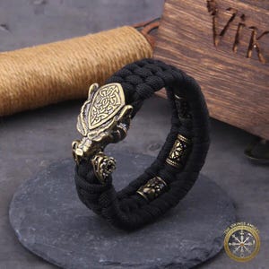 Puede incluir: Pulsera trenzada negra con una cabeza de drag&oacute;n vikingo de color bronce y cuentas decorativas. La pulsera se exhibe sobre una superficie de piedra gris oscuro, con una caja de madera y cuerda en el fondo. Una joya &uacute;nica.