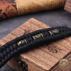 Puede incluir: Pulsera trenzada negra con cuentas decorativas de color bronce. Las cuentas presentan varios dise&ntilde;os de inspiraci&oacute;n vikinga, incluyendo una pata de oso y patrones geom&eacute;tricos. La pulsera se exhibe sobre una superficie de madera.