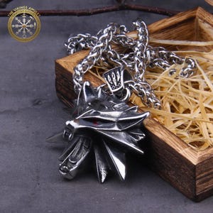 May include: A silver-colored pendant necklace featuring a wolf head with red eyes. The pendant is attached to a chain. The necklace is displayed in a wooden box with straw.