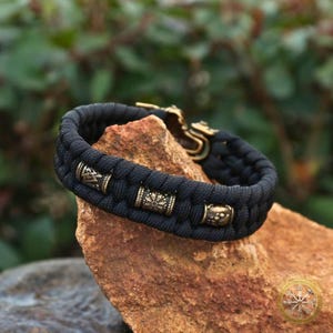 Puede incluir: Pulsera trenzada negra con cuentas decorativas y cierre de color bronce. La pulsera descansa sobre una roca marr&oacute;n, con un fondo verde borroso. Las cuentas tienen patrones intrincados.
