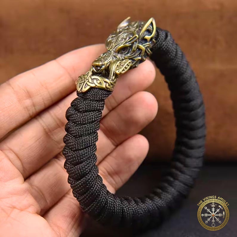 Puede incluir: Pulsera trenzada negra con un cierre de cabeza de lobo de color bronce. La cabeza de lobo presenta detalles intrincados. La pulsera es de color oscuro y parece estar hecha de un material tejido duradero. El logotipo de The Vikings Amulet es visible.