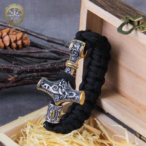 Puede incluir: Una pulsera trenzada negra con un colgante de martillo de Thor plateado y dorado. El martillo presenta detalles intrincados y se muestra dentro de una caja de madera. El logotipo de Vikings Amulet es visible.