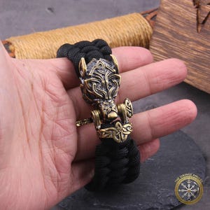 Puede incluir: Una pulsera trenzada negra con un dije de metal color bronce. El dije representa una cabeza de lobo estilizada y un dise&ntilde;o de martillo. La pulsera se sostiene en una mano, mostrando los intrincados detalles del metal.