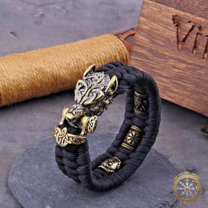 Puede incluir: Una pulsera trenzada negra con elementos decorativos de color bronce. La pulsera presenta un cierre detallado con cabeza de lobo y varias cuentas cil&iacute;ndricas con intrincados patrones. El dise&ntilde;o tiene un tema vikingo.