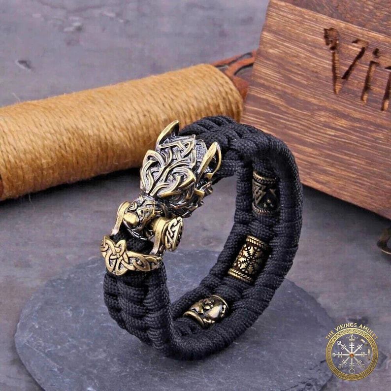 Puede incluir: Una pulsera trenzada negra con elementos decorativos de color bronce. La pulsera presenta un cierre detallado con cabeza de lobo y varias cuentas cil&iacute;ndricas con intrincados patrones. El dise&ntilde;o tiene un tema vikingo.
