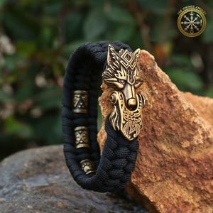 Puede incluir: Pulsera trenzada negra con un cierre de cabeza de lobo de bronce y tres cuentas de bronce. La cabeza de lobo presenta detalles intrincados. La pulsera se muestra sobre una roca, con el texto "THE VIKINGS AMULET" en un logotipo circular.