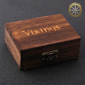 Puede incluir: Una peque&ntilde;a caja rectangular de madera con la palabra "Vikings" grabada en la parte superior. La caja est&aacute; te&ntilde;ida de color marr&oacute;n oscuro y tiene un cierre met&aacute;lico. El logotipo "The Vikings Amulet" est&aacute; en la esquina superior derecha.