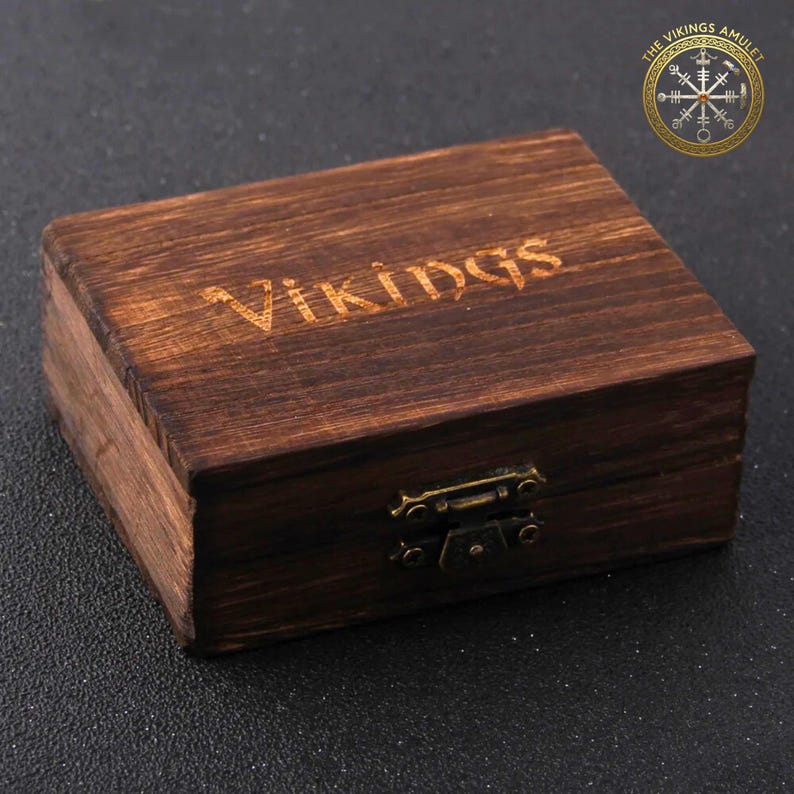 Puede incluir: Una peque&ntilde;a caja rectangular de madera con la palabra "Vikings" grabada en la parte superior. La caja est&aacute; te&ntilde;ida de color marr&oacute;n oscuro y tiene un cierre met&aacute;lico. El logotipo "The Vikings Amulet" est&aacute; en la esquina superior derecha.