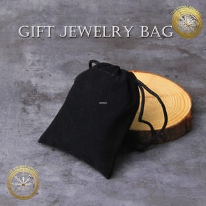 Puede incluir: Una bolsa de regalo negra con cord&oacute;n para joyas sobre una rodaja de madera. La bolsa est&aacute; hecha de un material suave y tiene un cierre de cord&oacute;n. El texto "GIFT JEWELRY BAG" se muestra encima de la bolsa.