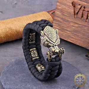 Puede incluir: Pulsera trenzada negra con cierre de cabeza de lobo color bronce y cuentas decorativas. La cabeza de lobo presenta un escudo con un dise&ntilde;o intrincado. Accesorio de tem&aacute;tica vikinga.