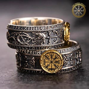 Puede incluir: Dos anillos plateados con intrincados diseños inspirados en los vikingos. Un anillo presenta un emblema dorado con un símbolo de rosa de los vientos. Los anillos están apilados, mostrando tallas detalladas de criaturas míticas y runas.