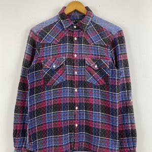 Könnte beinhalten: Ein langärmeliges kariertes Hemd in Lila, Blau, Schwarz und Grau mit zwei Brusttaschen und einem kontrastierenden blau karierten Joch. Das Hemd ist geknöpft und hängt an einem Holzbügel.