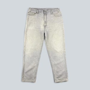May include: A pair of light gray denim jeans featuring a classic five-pocket design, a button and zipper fly closure, and belt loops along the waistband. The fabric has a faded, washed-out appearance with visible stitching along the seams.