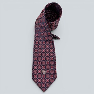 Puede incluir: Una corbata con fondo azul marino oscuro y un patrón repetitivo de pequeños y complejos motivos florales circulares en rojo y blanco. La parte inferior de la corbata muestra un pequeño logotipo bordado en hilo blanco cerca de la punta.