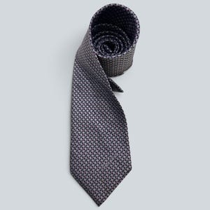 Puede incluir: Una corbata con un patrón geométrico repetitivo de pequeños cuadrados de color morado oscuro y gris claro. La corbata está enrollada en la parte superior, mostrando el forro interior, y se estrecha hacia un extremo puntiagudo.