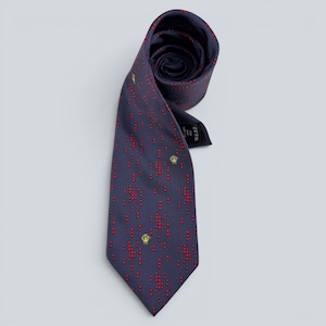 May include: A dark blue necktie featuring a pattern of small red dots arranged in vertical lines and interspersed with small gold lion head emblems. The tie is partially rolled at the top, revealing a black fabric label on the inner side of the blade.