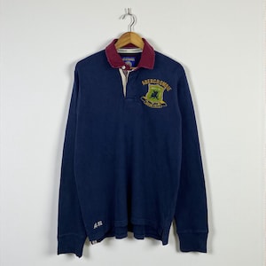 May include: Navy blue long-sleeve rugby shirt with a maroon collar and a button-up placket. Features an embroidered crest with the brand name "Abercrombie" and a small "A-92" logo on the sleeve. The shirt is hanging on a wooden hanger.