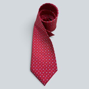 Puede incluir: Una corbata roja con un patrón repetitivo de pequeños motivos de aves en azul claro y blanco. La corbata está enrollada en la parte superior, mostrando la tela interior, y se extiende hacia abajo hasta una punta, con el estampado.