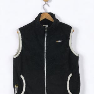 May include: Black fleece vest with a high collar and a full-length zipper. The vest has white trim along the edges and around the pockets. The National Geographic logo is visible on the front and side. The vest is hanging on a wooden hanger.