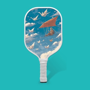 Peut inclure: Une raquette de pickleball avec une poignée blanche et un design unique. La face de la raquette présente une peinture d'une personne faisant du deltaplane dans un ciel bleu rempli d'oiseaux en origami et de nuages. La raquette est posée sur un fond turquoise.
