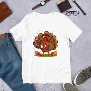Könnte beinhalten: Weißes T-Shirt mit einer Cartoon-Truthahn, der eine Sonnenbrille und ein Football-Trikot mit der Nummer 67 trägt. Der Truthahn hält einen Football, mit Herbstblättern um seine Füße. Ein lustiges, festliches Design.
