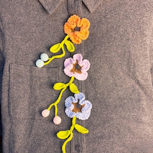 Könnte beinhalten: Ein anthrazitfarbenes Hemd mit Knopfleiste und drei gehäkelten Blumenapplikationen. Die Blumen sind orange, hellviolett und hellblau, jeweils mit einem dunkelbraunen Knopf in der Mitte. Grüne gehäkelte Stängel mit kleinen runden Knospen.