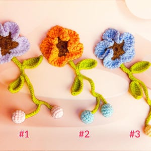 May include: Three crocheted flower bookmarks labelled #1, #2, and #3. Each features a five-petal flower in lavender, orange, or light blue with a brown centre, a green stem with two leaves, and two small round crocheted spheres at the bottom end.