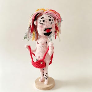 May include: A handcrafted crochet doll with pale skin, a black beard, and multicolored yarn hair in shades of red, yellow, and blue. The doll features embroidered facial details, pink cheeks, and holds a small red crocheted bag with a white flower.
