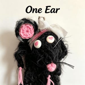 May include: A black crocheted plush toy with the text One Ear at the top. It features a single pink crocheted ear, white eyes with pink heart pupils, a pink crocheted nose, pink yarn tassels, and a white ribbon bow attached to the head.