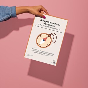 Może przedstawiać: Książka zatytułowana "Guía práctica de las emociones: cómo escucharlas y conectar" jest trzymana na różowym tle. Okładka zawiera ilustrację kompasu i tekst w języku hiszpańskim. Celem książki jest pomóc ludziom zrozumieć i połączyć się ze swoimi emocjami.