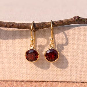 Puede incluir: Un par de pendientes colgantes dorados con piedras preciosas redondas de color rojo intenso. Los pendientes cuelgan de una pequeña rama curvada sobre un fondo neutro. Las piedras están engastadas en un bisel dorado.