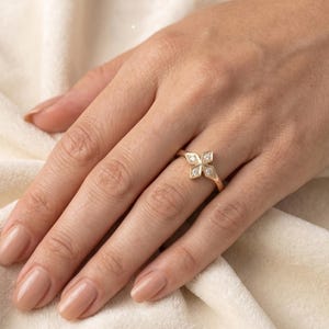 May include: A gold ring featuring a four-petal flower design with small, clear gemstones set in each petal. The ring is on a hand with neutral-toned nail polish, set against a soft, off-white background.