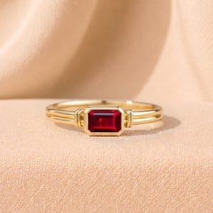 Op de afbeelding: Een gouden ring met een rechthoekige, dieprode edelsteen. De ring heeft een dubbelbandontwerp met decoratieve elementen aan weerszijden van de edelsteen. De achtergrond is een zachte, neutrale kleur.