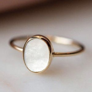 Könnte beinhalten: Ein goldener Ring mit einem ovalen, weißen Edelstein. Der Ring hat ein schmales Band und eine Lünettenfassung, die den Edelstein einfasst. Der Ring ist vor einem weichen, verschwommenen Hintergrund platziert, der das Design des Schmucks hervorhebt.