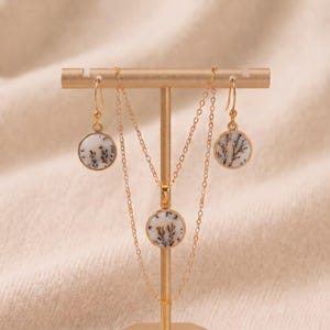 May include: A gold jewellery set consisting of a necklace and two matching drop earrings. Each piece features a circular pendant with a white stone inlay displaying dark, branching, tree-like dendrite inclusions set in a thin gold frame.