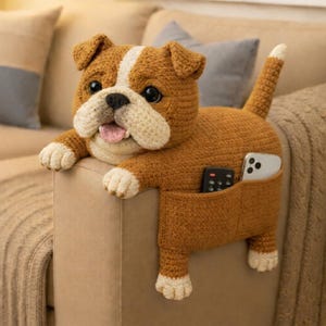 May include: Crocheted bulldog-shaped couch armrest organizer in shades of brown, cream, and white. The dog has a pocket for storing a remote control and a phone. The dog's tongue is pink.