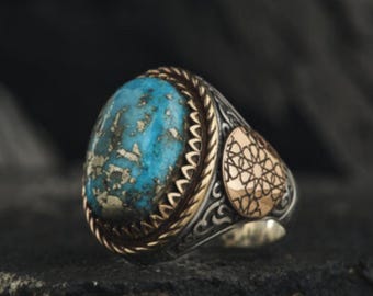 May include: A silver ring with a large oval turquoise stone. The ring has gold accents around the stone and on the side, featuring an intricate pattern. The ring is set against a dark, textured background.