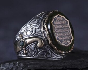 May include: Silver ring with an oval green stone and gold accents. The ring features intricate scrollwork and a gold band. The green stone has gold text. The ring is displayed on a dark, textured surface.