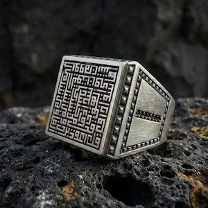 Op de afbeelding: Een zilverkleurige ring met een vierkant oppervlak met ingewikkelde zwarte kalligrafie. De ring heeft een rechthoekige band met een getextureerd oppervlak en kleine, donkere accenten. De ring wordt getoond op een donker, getextureerd oppervlak.