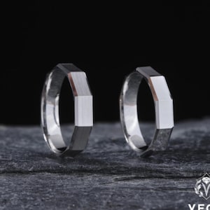 May include: Two silver rings with geometric designs. Each ring features a rectangular center section flanked by angled facets, creating a modern, angular look. The rings are displayed on a dark, textured surface.