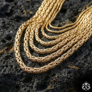 Può includere: Primo piano di più collane a catena bizantina color oro. Le catene variano di spessore e sono esposte su una superficie scura e strutturata. Le collane sono finemente collegate, mostrando un classico design di gioielli.