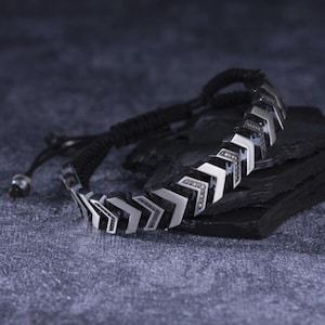 May include: A silver and black bracelet with an arrow design. The bracelet features a series of chevron-shaped links, connected by a black cord. The bracelet is displayed on a dark, textured surface, creating a contrast with the silver elements.