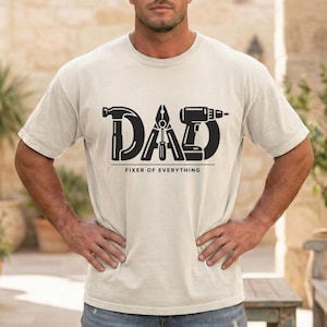 Könnte beinhalten: Ein hellbeige T-Shirt mit dem Wort "DAD" in schwarzen Buchstaben, das Werkzeuggrafiken enthält. Hammer, Zange, Schraubendreher und Bohrmaschine bilden die Buchstaben. Darunter steht der Text "FIXER OF EVERYTHING".